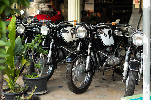 Parked restored old BMW R models motorbikes in Bangkok are standing outside of garage shop on sidewalk in Nak Niwat. Inside of garage is a man