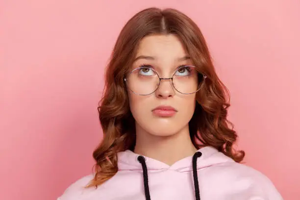 Portrait of puzzled female brunette teen in round eyeglasses looking up, thinking about information, doubting about choice. Indoor studio shot, isolated on pink background