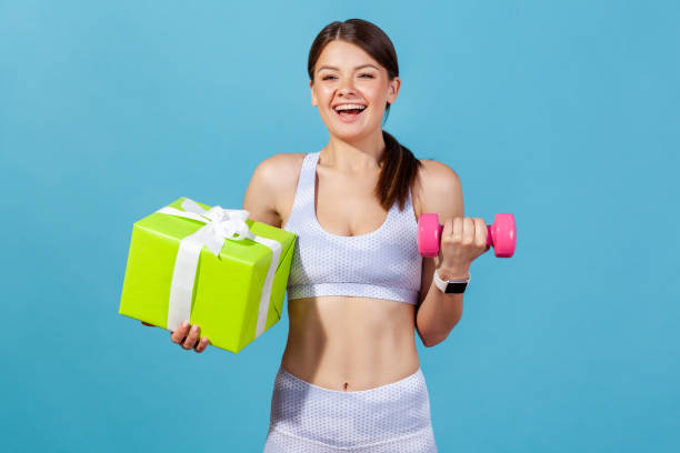 smiling positive woman in white sportswear holding pink dumbbell and big green gift box, receiving gym membership on holidays - muscular build bicep women female imagens e fotografias de stock