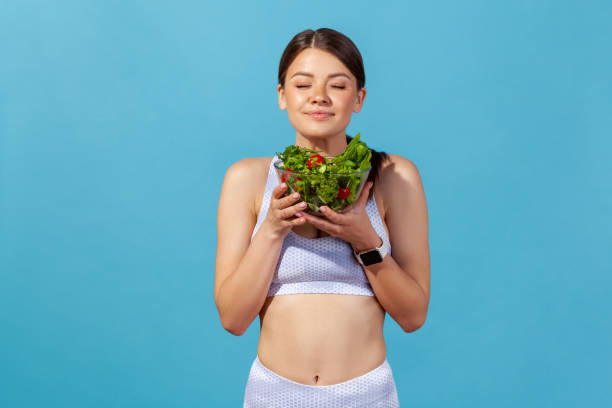 Athletic slim woman in white sports top closing eyes enjoying aroma of fresh vegetable salad in big bowl, healthy nutrition, detox diet Athletic slim woman in white sports top closing eyes enjoying aroma of fresh vegetable salad in big bowl, healthy nutrition, detox diet. Indoor studio shot isolated on blue background eating body building muscular build vegetable stock pictures, royalty-free photos & images