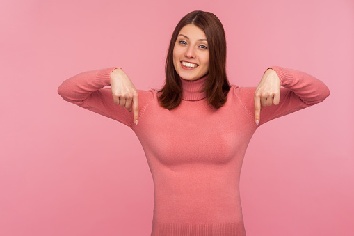 Cheerful positive woman in pink sweater pointing fingers down showing space for your advertisement, looking at camera with toothy smile. Indoor studio shot isolated on pink background