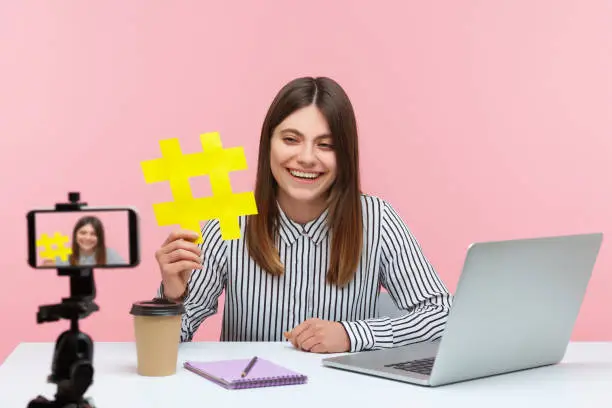 Photo of Smiling attractive woman blogger showing big yellow hashtag symbol at smartphone camera, sharing viral content, tagged message