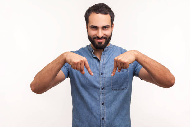 Satisfied successful man with beard pointing fingers down paying attention at advertising area, showing freespace, looking at camera with smile Satisfied successful man with beard pointing fingers down paying attention at advertising area, showing freespace, looking at camera with smile. Indoor studio shot isolated on white background man hand down stock pictures, royalty-free photos & images