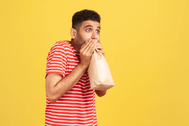 istock Profile portrait scared man in striped red t-shirt exhaling and inhaling into package, using paper bag to improve well-being, overcoming stress fear 1291590761