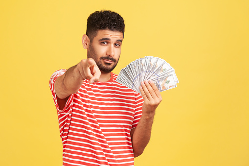 Self confident rich man with beard in striped t-shirt holding fan of dollars and pointing finger at camera with serious expression, easy profit. Indoor studio shot isolated on yellow background