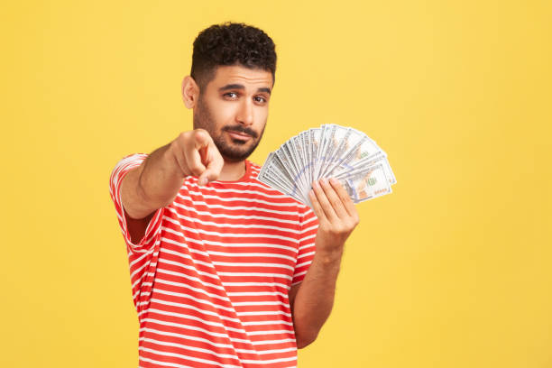 self confident rich man with beard in striped t-shirt holding fan of dollars and pointing finger at camera with serious expression, easy profit. - iranian currency imagens e fotografias de stock