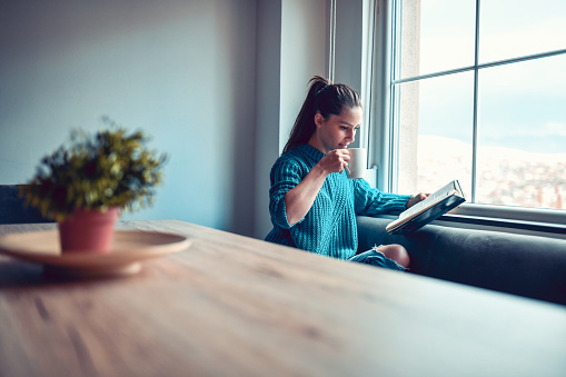 Cute Female Sitting By The Window, Drinking Coffee And Reading A Book