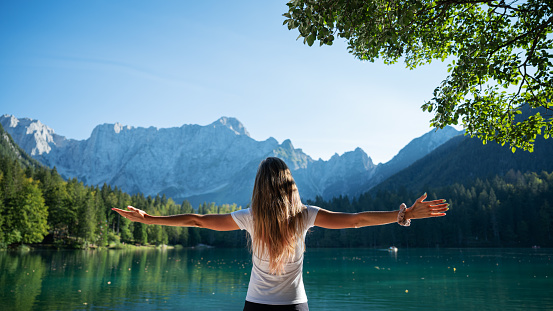 View from behind of a young woman with long brown hair standing by a beautiful green lake with her arms spread widely, lit by the sun.
