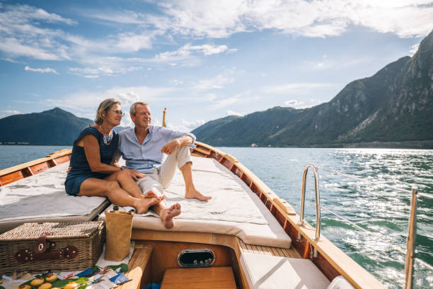 Mature couple relax on sailboat moving through Lake Lugano Mountain range behind is lit by the morning light luxury lifestyle stock pictures, royalty-free photos & images