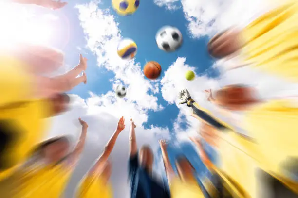 Group of happy sports team with coach throwing team sports balls. Summer sky with clouds in the background. Children playing team sports outdoor; soccer, basketball, volleyball, handball, tennis ball