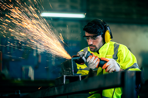 Caucasian male worker in a safty uniform wear welders leathers,Electric wheel grinding on steel structure in factor in the garage, orange bokeh sparks fly to the sides