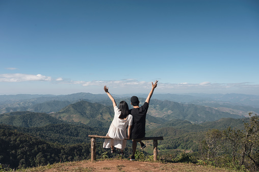 Young asian couple sitting raised arms on top of hill in countryside on vacation