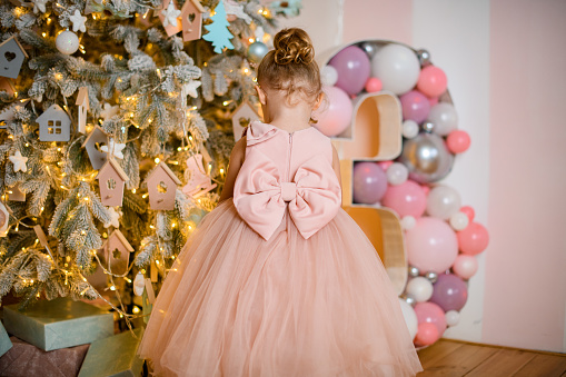 back view of little girl in beautiful pink dress with tied bow at the belt standing near christmas tree