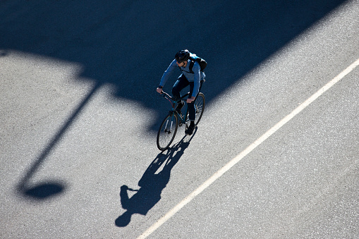 A male bicycle commuter rides a city street on his way to work. He is wearing a bicycle helmet, wears casual clothes, and carries a bicycle messenger style backpack. He rides a fixed-gear or one-speed bicycle.