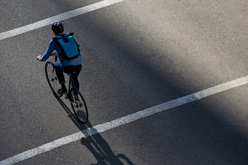A male bicycle commuter rides a city street on his way to work. He is wearing a bicycle helmet, wears casual clothes, and carries a bicycle messenger style backpack. He rides a fixed-gear or one-speed bicycle.