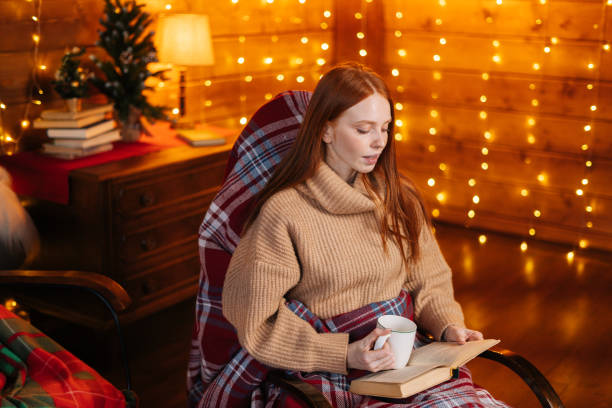 istock Charming young woman holding cup with hot coffee and reading book while sitting at rocking armchair 1291640809