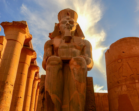 Upward angle of giant seated figure in a temple in Luxor with blue sky and feathery clouds in the background.