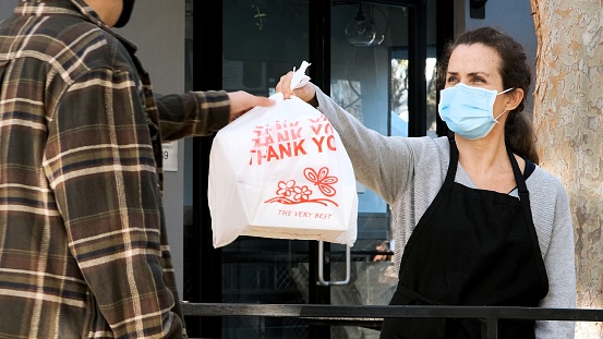 Caucasian mature restaurant female worker delivering a bag with meal at a restaurant curbside pickup wearing a protective face mask