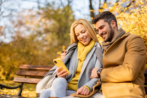 Romantic couple sitting on a park bench on a beautiful autumn day  and using a smart phone.