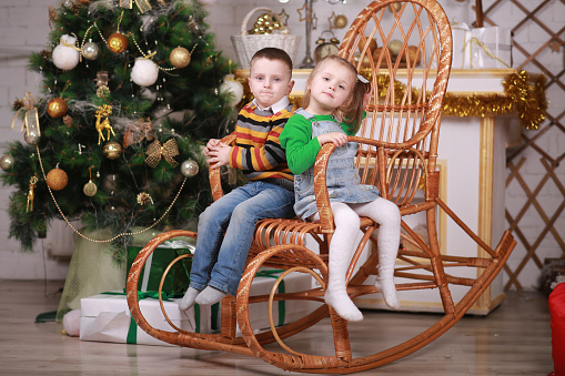 cute little girl and boy sitting in rocking chair near christmas tree.
