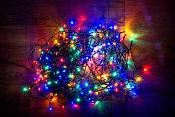 A pile of tangled Christmas tree lights. The brightly colour LED bulbs make a colourful display, having been plugged in and turned on to show a variety of glowing colours on a long, black wire. The red, blue, green, yellow, red and pink lights are heaped on a wooden floor, in preparation for being untangled, which is a frustrating task that needs completing before the lights are used to decorate a household Christmas tree.