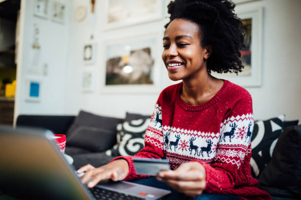 Young happy African American woman shopping online Beautiful African American woman shopping online from her cozy home office during winter holidays, holding a credit card while purchasing something online using a laptop holiday shopping stock pictures, royalty-free photos & images