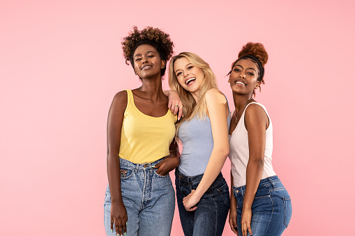 Fashion photo of three beautiful multiethnic women smiling and looking at camera, posing over pink pastel background. Beauty lifestyle and multiracial friendship concept.