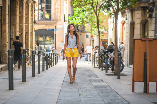 Young Sightseer Enjoying Barcelona Side Street in Summer Full length front view of female vacationer in mid 20s wearing casual summer clothing and  looking at view while approaching camera on Barcelona side street. approaching stock pictures, royalty-free photos & images