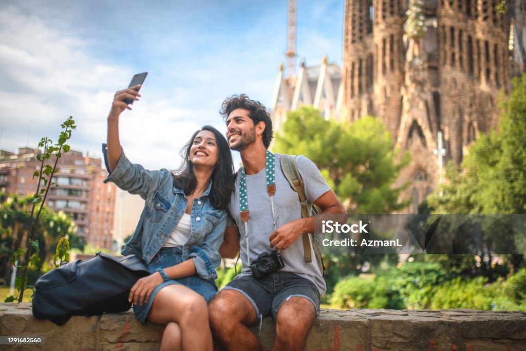 Young Couple Taking Break from Sightseeing for Selfie Male and female travelers sitting on wall in public park near Sagrada Familia in Barcelona and taking selfie on sunny summer day. Couple - Relationship Stock Photo