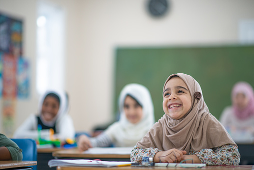 A happy and smiling Muslim school student is sitting at her desk in her classroom. In the background, her other female classmates can be seen wearing a hijab as well.
