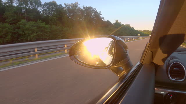 CLOSE UP: Blinding golden sunbeams are reflected in the side mirror of a car.