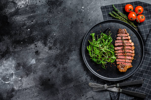 istock Cooked grilled strip loin steak, marbled beef meat with arugula. Black background. Top view. Copy space. 1291718321