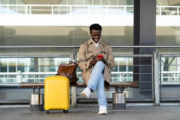 schwarzer reisender, mann, der auf bank im flughafenterminal mit mobiltelefon sitzt und auf öffentliche verkehrsmittel wartet - airport lounge airport one person bag stock-fotos und bilder