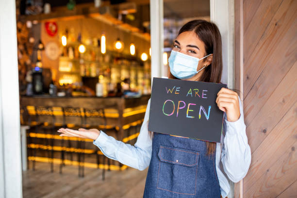 istock Happy female waitress with protective face mask holding open sign while standing at cafe or restaurant doorway, open again after lock down due to outbreak of coronavirus covid-19 1291734047