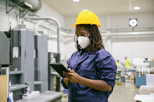 African American female factory worker in mask holding tablet. Focused content skilled woman working on plant and wearing protective helmet and uniform. Manufacture and digital technology concept