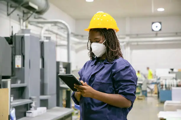 Photo of African American female factory worker in mask holding tablet