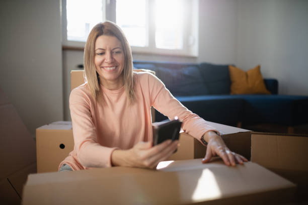 istock Smiling woman sitting amid cardboard boxes and using phone 1291737866