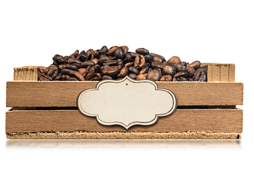 Closeup of roasted coffee beans in a small wooden crate with empty label, isolated on white background with reflections.