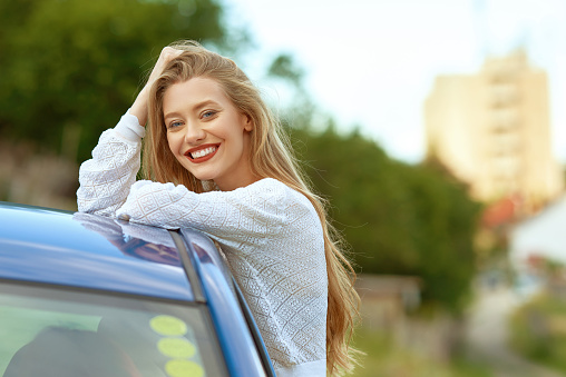 Blond woman with toothy smile leaning on car rooftop in nature looking at the camera