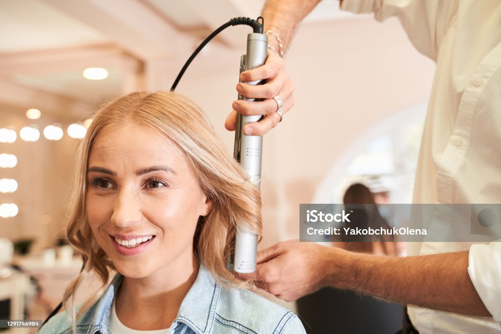 Woman enjoying of the beauty procedures Going for big curls. Young beautiful woman enjoying of the beauty procedures with her hairdresser while sitting in the hair salon and getting her hair done with hair iron Curling Tongs Stock Photo