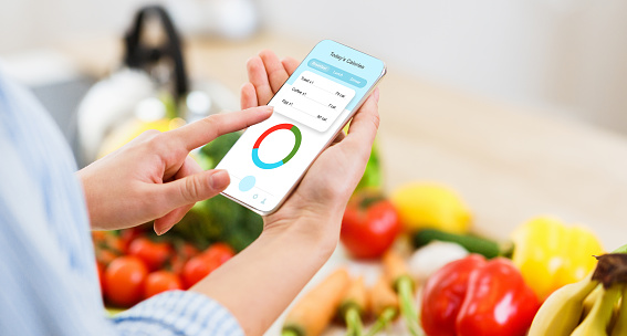 Woman using calorie counter application on her smartphone while cooking healthy lunch in kitchen. Fresh tasty vegetables and fruits lying on table on background, creative collage, panorama