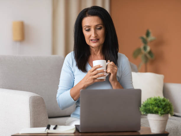 istock Mature woman sitting on sofa drinking tea working on computer 1291754984