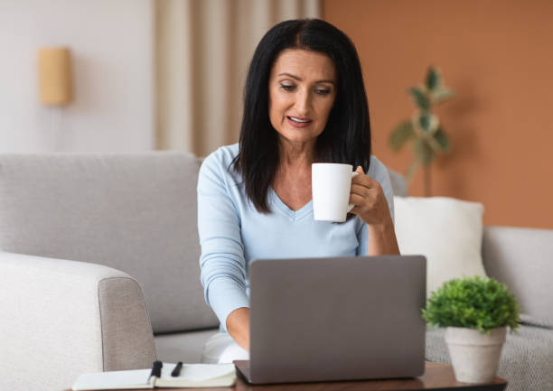 istock Mature woman sitting on sofa drinking coffe working on computer 1291755073