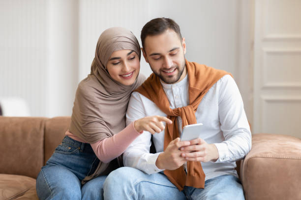 istock Muslim Couple Using Mobile Phone Sitting On Sofa At Home 1291755181