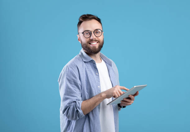 istock Positive young guy in casual wear using tablet computer over blue studio background 1291759690