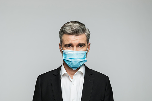 Portrait of worried mature businessman wearing N95 face mask looking at camera. Studio shot, grey background.