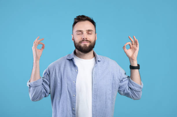 istock Stress management. Peaceful young guy meditating with closed eyes, making gyan mudra with his hands on blue background 1291764984