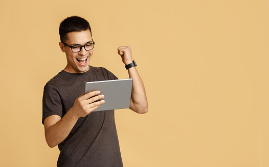 Online games, cashback and emotions of winning. Happy young handsome male in glasses looks in digital tablet and expresses success, isolated on light sand background, free space, studio shot