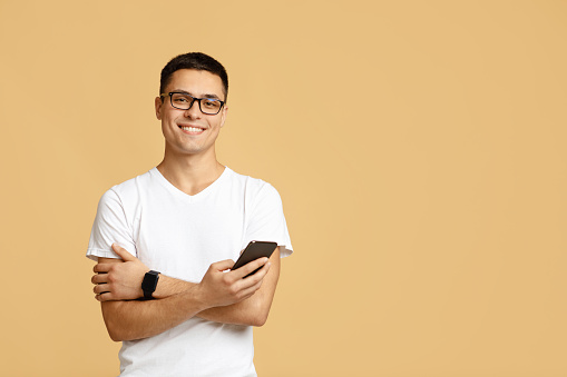 Social media, app, modern communication and lockdown. Attractive cute zoomer student in glasses, white t-shirt with smart watch hold smartphone and texting message, isolated on light sand background