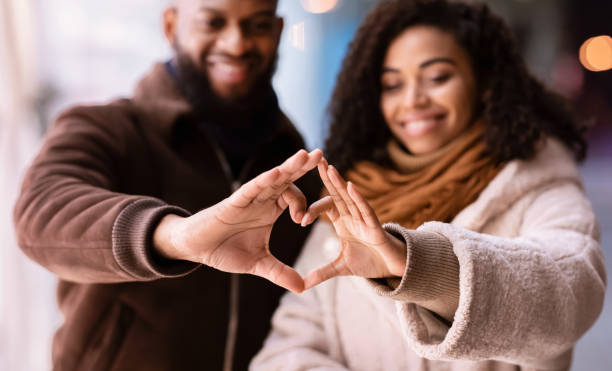 Celebrating Valentine's Day. Close up of smiling loving black man and woman making heart shape with fingers walking together in the evening, looking at hands, selective focus, blurred background